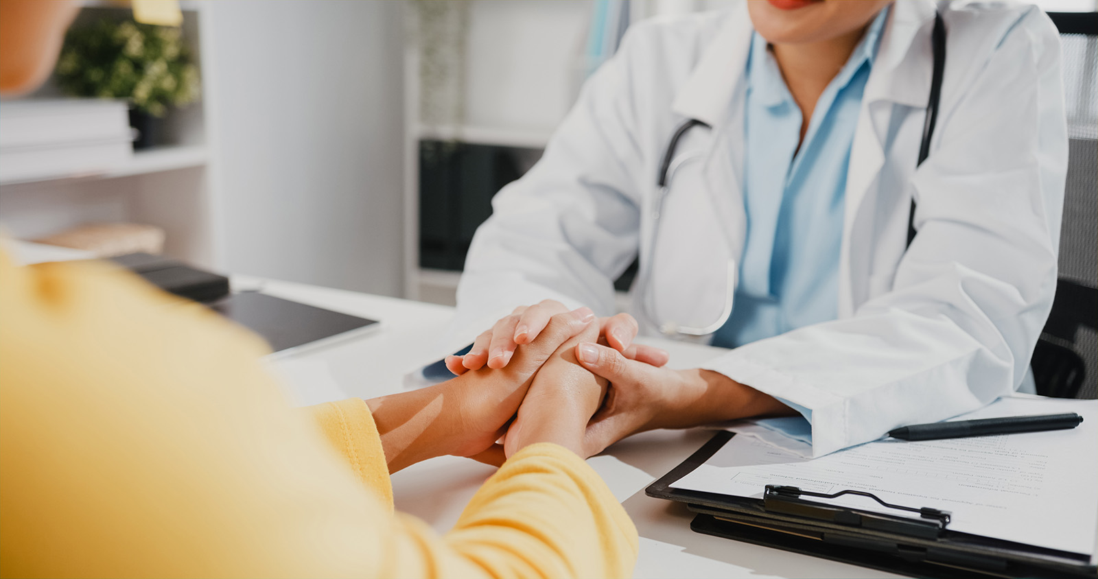 Close up of young Asia female doctor in white medical uniform talk discuss touching girl patient shoulder at meeting sitting at desk in health clinic or hospital office